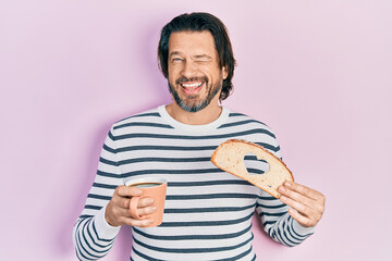 Middle age caucasian man holding bread loaf with heart shape drinking coffee winking looking at the...