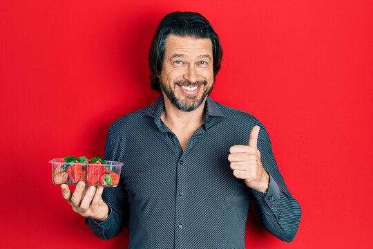 Middle age caucasian man holding strawberries smiling happy and positive, thumb up doing excellent and approval sign