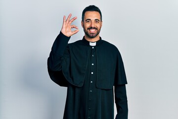 Handsome hispanic man with beard wearing catholic priest robe smiling positive doing ok sign with...