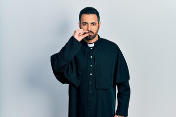 Handsome hispanic man with beard wearing catholic priest robe mouth and lips shut as zip with...