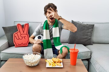 Young hispanic man football hooligan cheering game at home peeking in shock covering face and eyes with hand, looking through fingers with embarrassed expression.