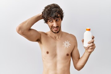 Young hispanic man standing shirtless holding sunscreen lotion confuse and wondering about...