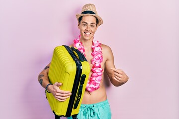 Young hispanic man wearing summer hat and holding cabin bag smiling happy pointing with hand and finger