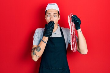 Young hispanic man wearing cook apron and holding meat covering mouth with hand, shocked and afraid for mistake. surprised expression