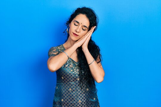 Young woman wearing bindi and traditional kurta dress sleeping tired dreaming and posing with hands together while smiling with closed eyes.