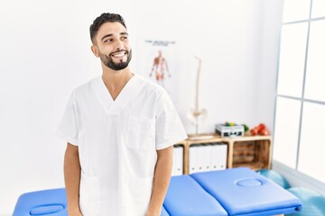 Young handsome man with beard working at pain recovery clinic looking away to side with smile on face, natural expression. laughing confident.