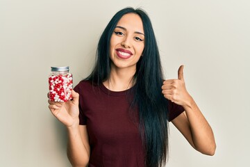 Young hispanic girl holding pills smiling happy and positive, thumb up doing excellent and approval sign