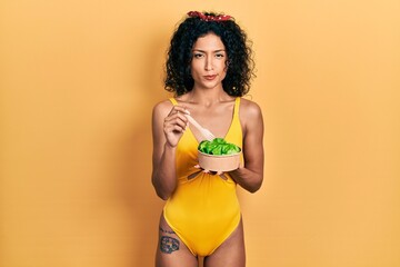Young latin girl wearing swimsuit eating salad skeptic and nervous, frowning upset because of...