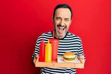 Middle age hispanic man eating a tasty classic burger and soda smiling and laughing hard out loud because funny crazy joke.