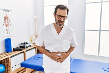 Middle age man with beard working at pain recovery clinic with hand on stomach because indigestion, painful illness feeling unwell. ache concept.