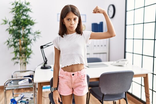 Young hispanic girl standing at pediatrician clinic strong person showing arm muscle, confident and proud of power