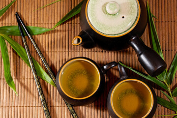 Japanese teapot and cups with green tea on a bamboo mat with bamboo leaves and chopsticks top down flat lay view.