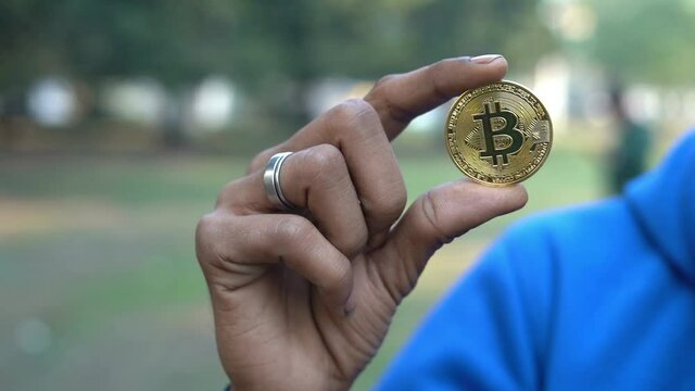Shot of an Asian Hand holding cryptocurrency bitcoin coin outdoors in park background. Shiny golden BTC crypto coin.