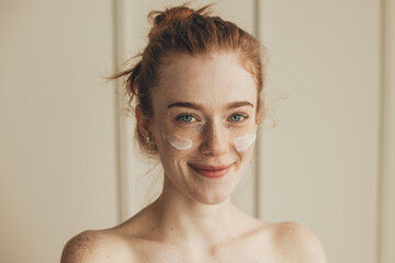 Close up portrait of a woman with red hair and freckles applying cream on her face. Facial...