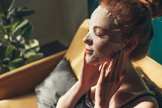 Redheaded woman applying facial mask on her face while sitting on sofa at home. Close up portrait. Beauty skin care. Rejuvenation treatment. Facial skin