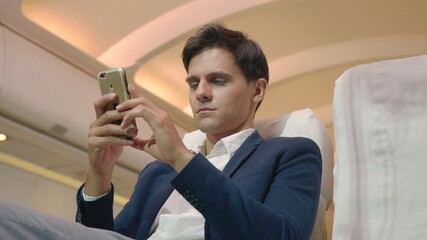 Businessman sitting inside plane watching video on mobile phone during his business trip, Young passenger man using smartphone on airplane while traveling for safe flight