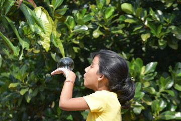 A girl kid is playing with soap water and making large bubbles wearing yellow shirt selectively focused green blurr background