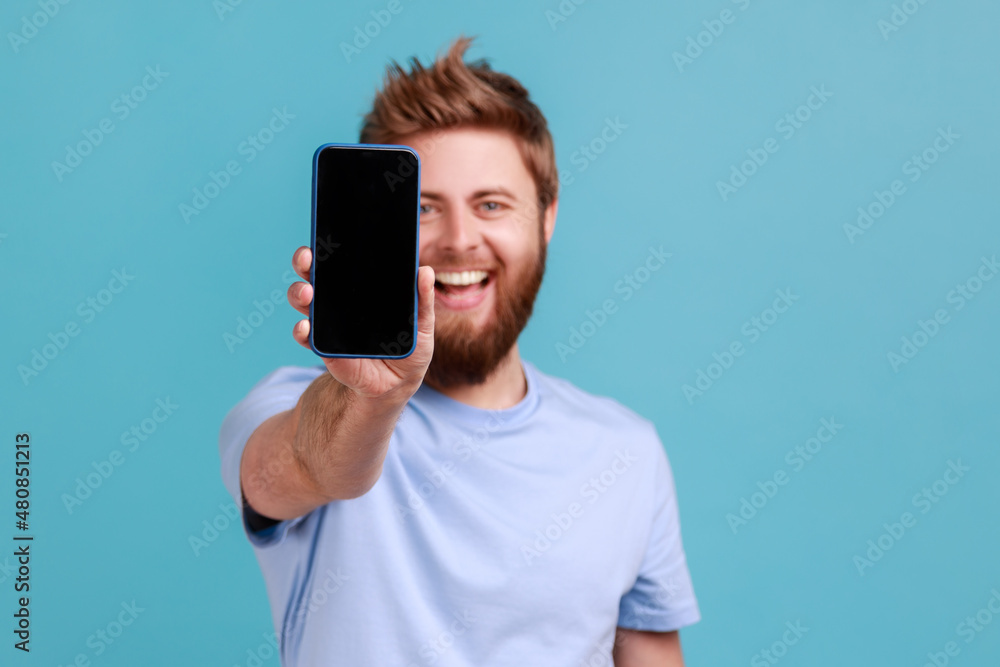 Wall mural portrait of bearded man holding out mobile phone and looking at camera with toothy smile and positiv