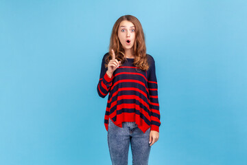 Excited young woman wearing striped casual style sweater standing raised finger up, having sudden idea, looking at camera with open mouth. Indoor studio shot isolated on blue background.