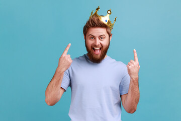 Portrait of excited amazed bearded man pointing fingers on golden crown on his head, showing her leadership qualities, concept of authority. Indoor studio shot isolated on blue background.