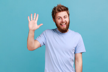 Portrait of positive bearded man greeting you rising hand and waving, saying hi, glad to see you, looking at camera with toothy smile. Indoor studio shot isolated on blue background. © khosrork