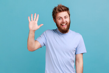 Portrait of positive bearded man greeting you rising hand and waving, saying hi, glad to see you,...