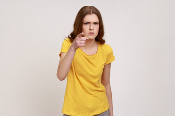 Hey you! Portrait of angry strict woman of young age in casual yellow T- shirt noticing and pointing finger to camera, accusing with serious bossy face. Indoor studio shot isolated on gray background.