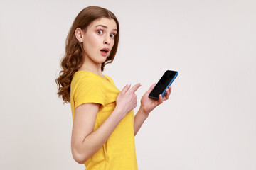 Naklejka premium Surprised brown haired teenager girl in yellow casual t-shirt pointing finger at smartphone in her hands, shocked with new phone features. Indoor studio shot isolated on gray background.