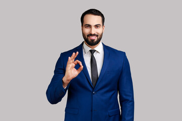 Portrait of delighted bearded man standing, looking at camera showing Ok sign gesture, expressing positive emotions, wearing official style suit. Indoor studio shot isolated on gray background.