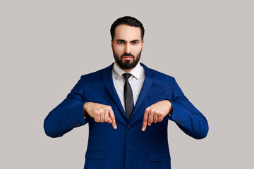 Serious bearded man pointing fingers down, demanding approach to him immediately, having control over situation, wearing official style suit. Indoor studio shot isolated on gray background.