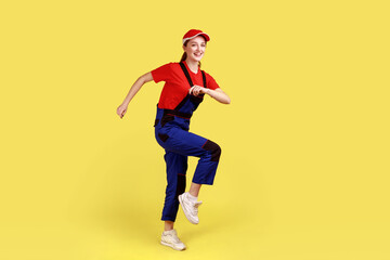 Fototapeta na wymiar Full length portrait of optimistic positive worker woman marching, posing on one leg, going to do her work, wearing overalls and red cap. Indoor studio shot isolated on yellow background.