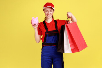 Portrait of happy courier woman standing with shopping bags in hands and piggybank, cashback, looking at camera, wearing overalls. Indoor studio shot isolated on yellow background.