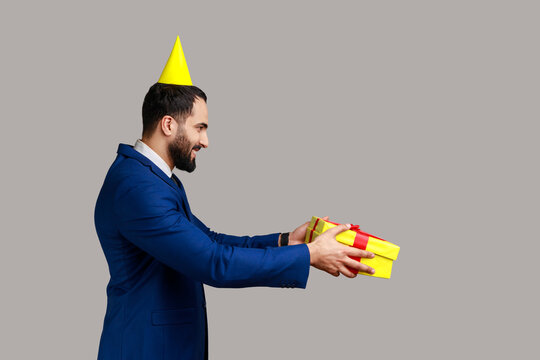 Side view of positive man with festive mood standing in party cone and giving present box, celebrating friend's birthday, wearing official style suit. Indoor studio shot isolated on gray background.