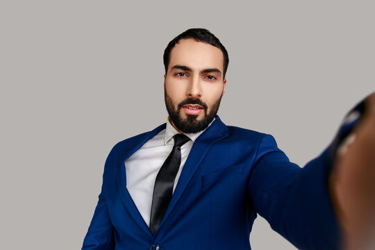 Portrait of handsome bearded man taking selfie, looking at camera with relaxed optimistic calm emotions, POV, wearing official style suit. Indoor studio shot isolated on gray background.