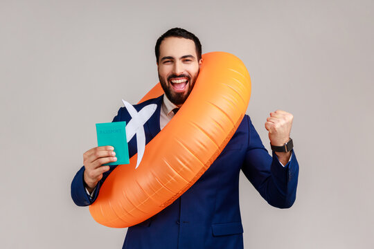Happy bearded businessman holding orange rubber ring, paper plane and passport, clenching fist, long awaited vacation, wearing official style suit. Indoor studio shot isolated on gray background.