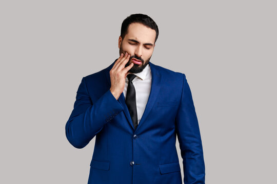 Unhealthy bearded man wincing in pain and touching sore cheek, suffering unbearable toothache, gum disease, wearing official style suit. Indoor studio shot isolated on gray background.