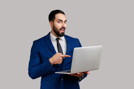 Astonished bearded man pointing laptop screen and looking at camera with shocked expression, open mouth, wearing official style suit. Indoor studio shot isolated on gray background.