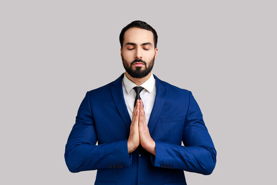 Portrait of calm handsome bearded businessman standing in yoga pose and try to relaxing, keeps palms together, wearing official style suit. Indoor studio shot isolated on gray background.