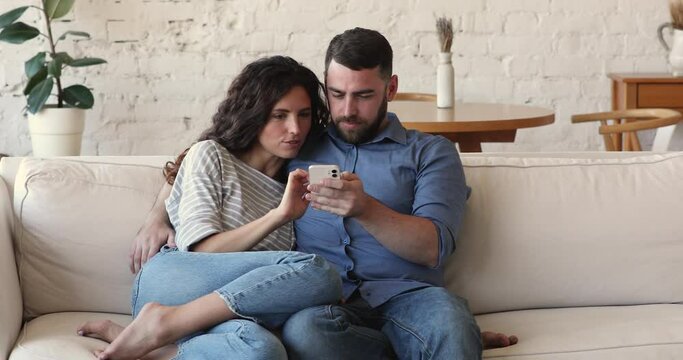 Attractive couple sit on sofa use cellphone, discuss purchase, enjoy new mobile app, consider order online. Internet connection, modern wireless tech usage, e-commerce retail services users concept