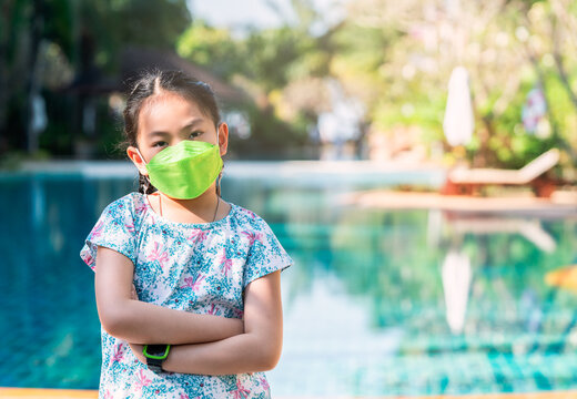 Portrait Asian child girl wearing a face mask, arms on the chest and standing beside swimming pool at a resort, concept for traveling in the era of covid-19 in Thailand. Day light image in a morning.