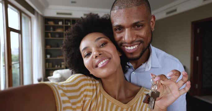 Excited African American couple modern apartment renters tenants hug hold keys shoot video on phone. Webcam portrait of overjoyed young husband wife share great news of getting mortgage or buying flat