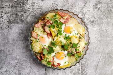Potatoe casserole with sausages, cheese and eggs in a glass baking dish on a dark grey background. Top view, flat lay.