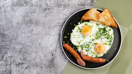 Fried eggs with green onions, sausages and toast on a round plate on a dark grey background. Top view, flat lay