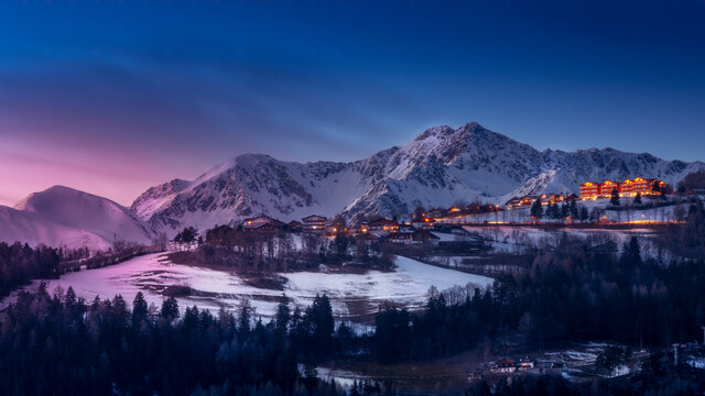 Scenic view of a town with hotels against blue sky and snowcapped mountains at sunset in winter. Snow and forest in the foreground