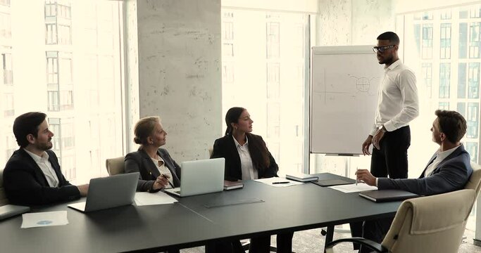 African coach or presenter speak at meeting, gives business presentation on flip chart for corporate staff. Businesspeople professional team take part in seminar in office board room. Training concept