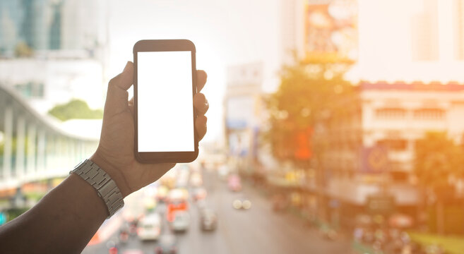 Hand holding black smartphone with white screen mockup and city blurred background with copy space , smartphone frameless application design concept.