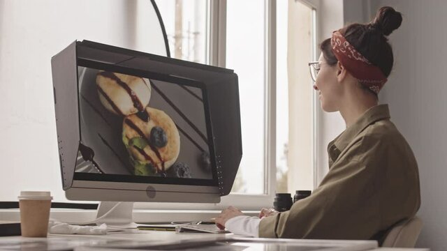Side-view medium shot of young female photographer editing food picture of delicious cheese pancakes on computer monitor while working in office