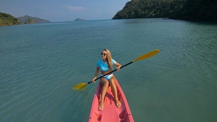 Pretty sportive blonde lady in stylish swimsuit sails on pink plastic canoe along azure ocean bay at exotic resort.