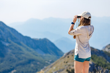Young woman watching mountain landscape through binoculars at the view spot on tourist trail.