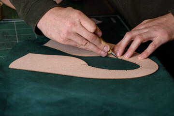 Shoemaker hands holding craft knife cutting green suede leather for handmade shoes on his workplace...